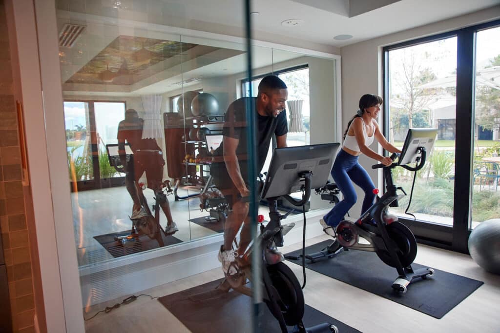 Two people are exercising on stationary bikes in a modern gym with large windows and mirrors, with natural light coming in.