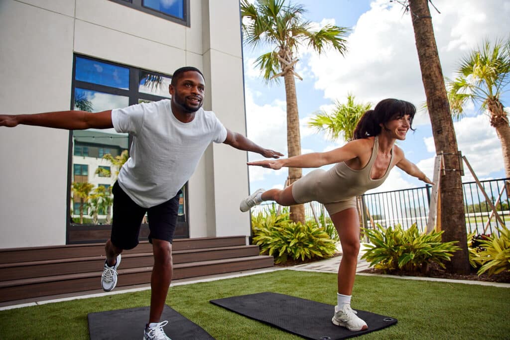 A man and woman practice balancing yoga poses on mats outdoors, with palm trees and a building in the background.