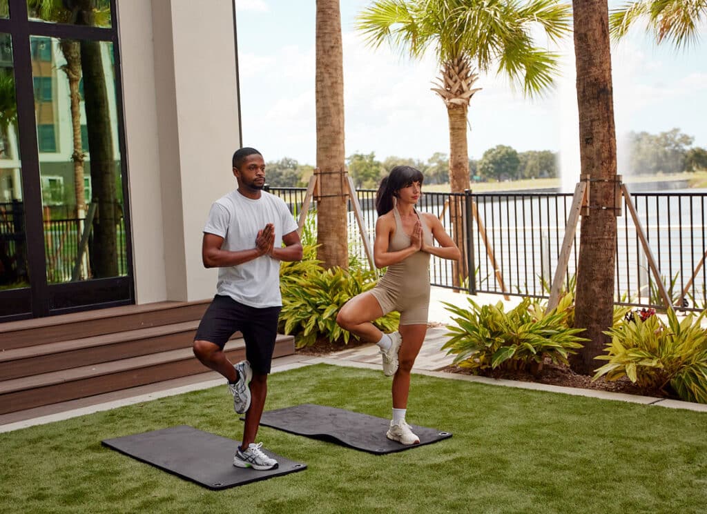 Two people practice yoga outdoors on mats, standing in tree pose near a fence with palm trees and a lake in the background.
