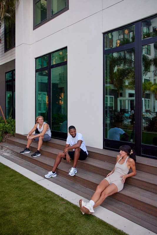 Three people in athletic wear sit and rest on outdoor wooden steps in front of a modern building with large windows.