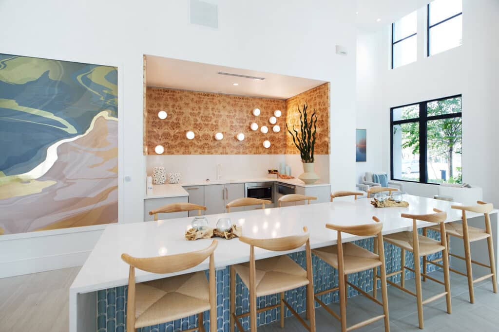 Modern kitchen and dining area with a long white island, wooden chairs, wall art, decorative lighting, and large windows letting in natural light.