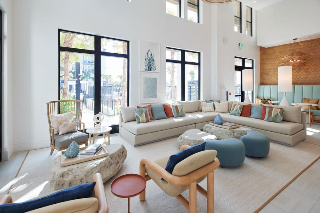 Modern, airy living room with large beige sectional sofa, colorful throw pillows, light wood chairs, stone coffee tables, and large windows letting in natural light.