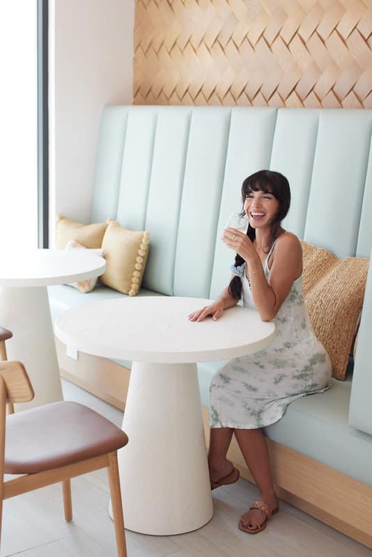 A woman in a light dress sits at a white table in a modern cafe, smiling and holding a glass of water.