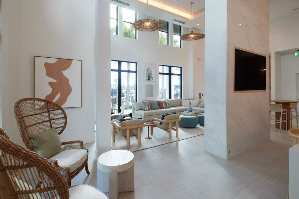 Modern living room with large windows, neutral tones, abstract art, rattan chairs, a sectional sofa, and a mounted TV on a marble wall. Natural light fills the space.