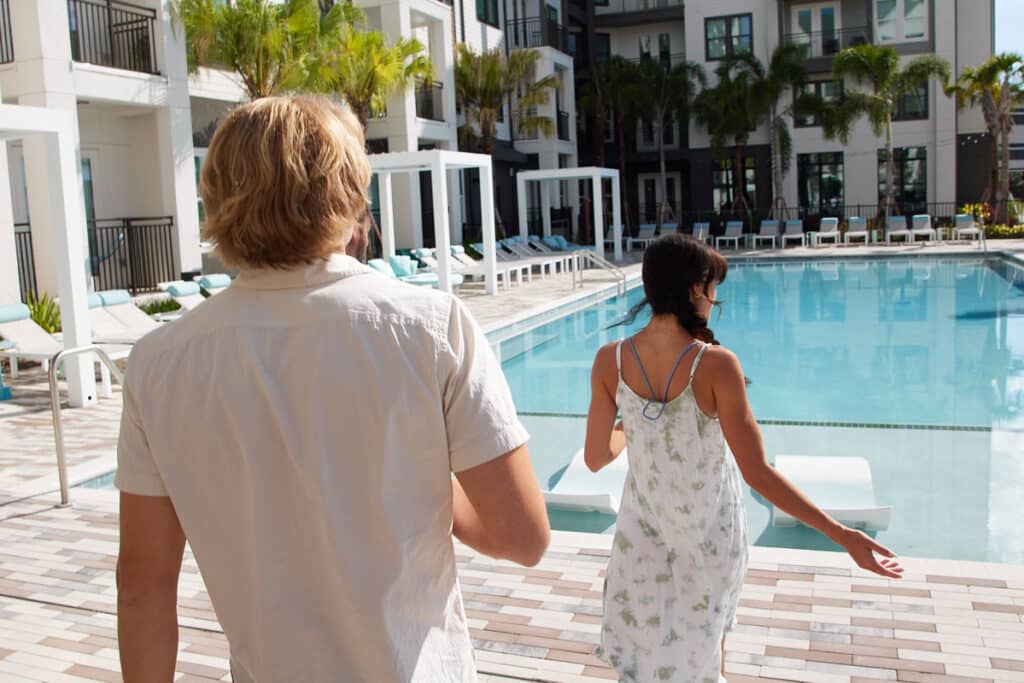 A man and a woman walk along the edge of an outdoor swimming pool at a modern apartment complex with palm trees and lounge chairs.