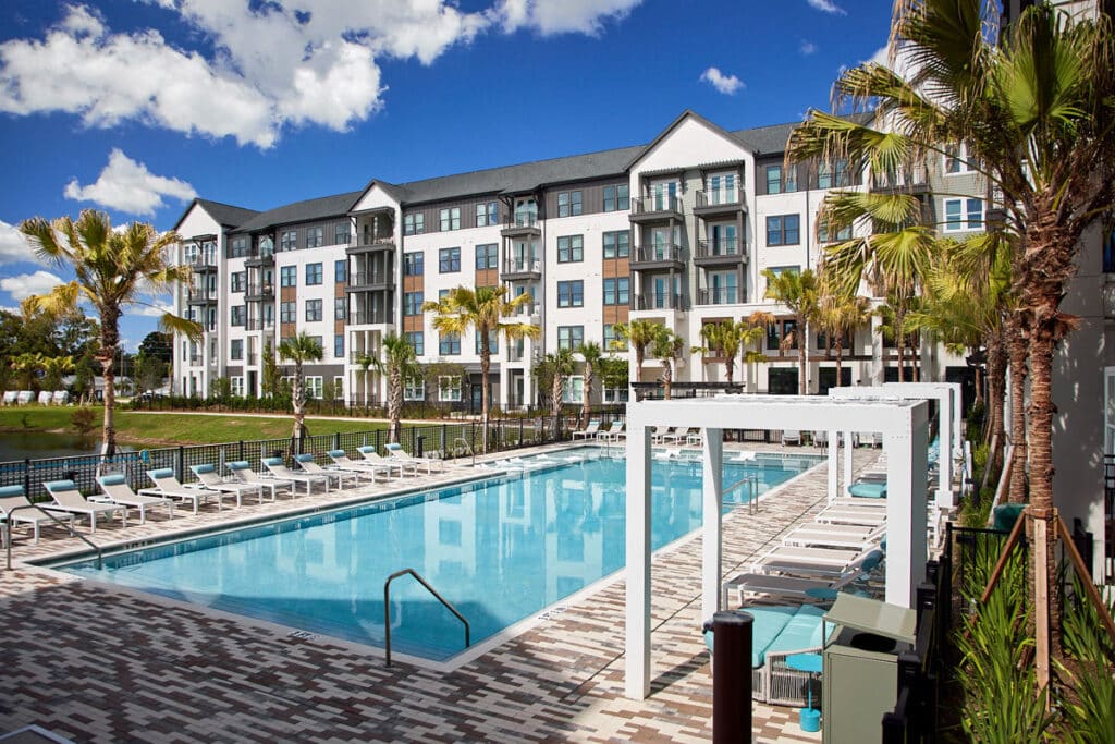 Rectangular outdoor pool surrounded by lounge chairs and palm trees, with a modern four-story apartment building in the background under a blue sky.