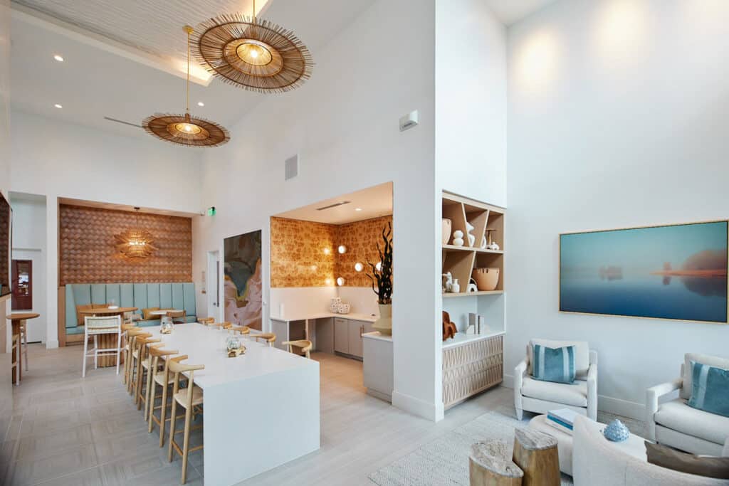 Modern, airy lounge with white walls, large windows, wooden furniture, a central table with stools, woven light fixtures, and decorative shelves.