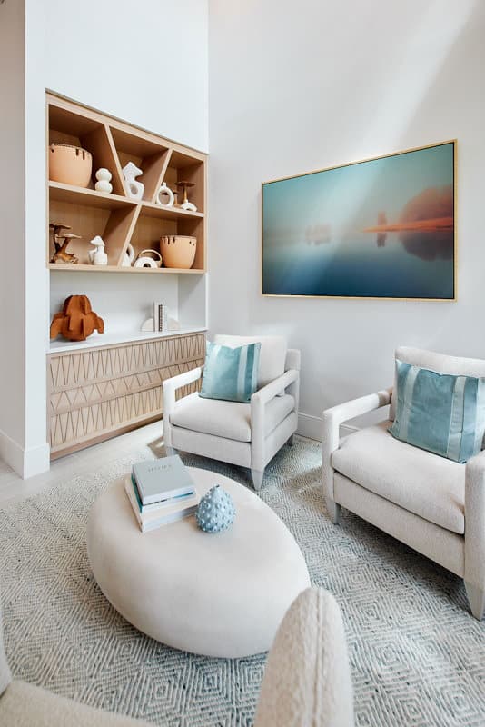 A modern living room with two white armchairs, a round ottoman, built-in shelving with ceramics, a textured rug, and a landscape artwork on the wall.