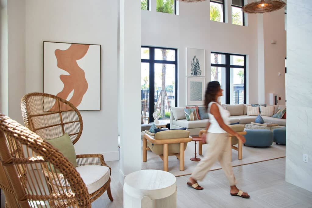 A woman walks through a modern, airy living room with large windows, neutral-toned furniture, woven chairs, and abstract artwork on the walls.