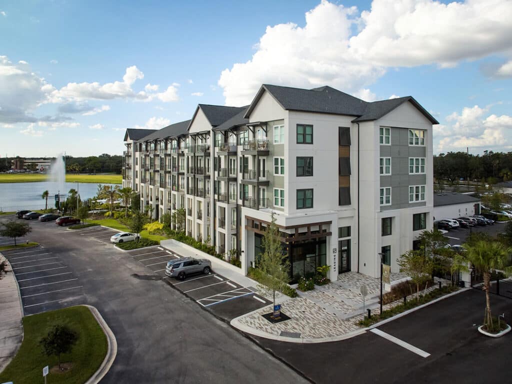 A four-story modern apartment building with balconies, surrounded by a parking lot, landscaping, and a lake with a fountain in the background under a partly cloudy sky.