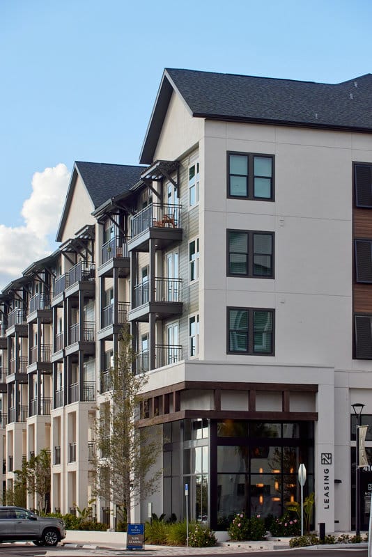 Four-story modern apartment building with balconies and large windows, featuring a street-level leasing office and a small tree in front.