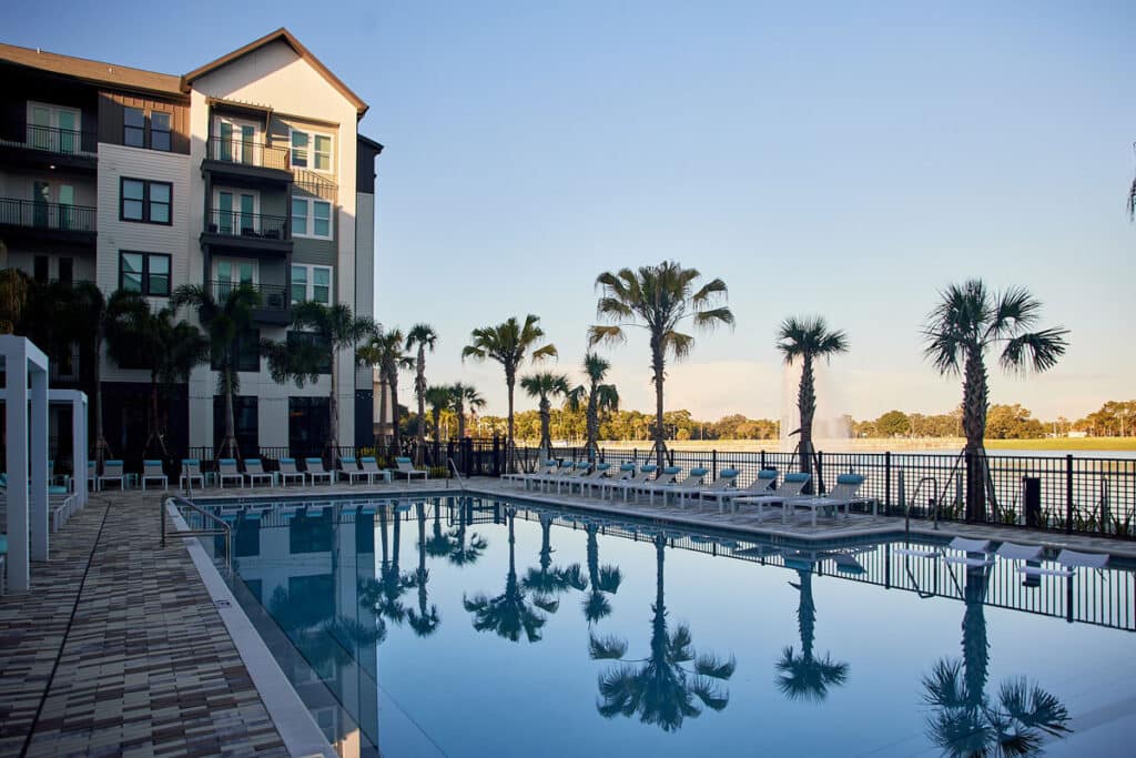 A large outdoor swimming pool is surrounded by lounge chairs, palm trees, and a multi-story building, with a lake visible in the background.