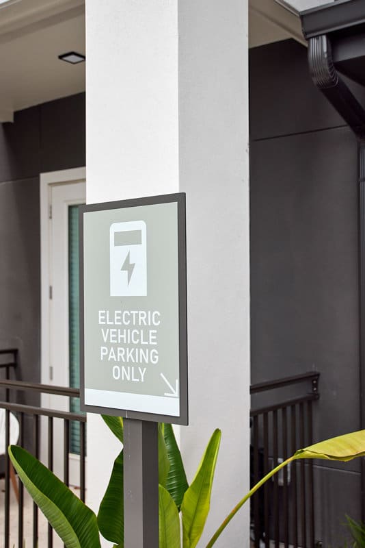 A sign reads "Electric Vehicle Parking Only" with a charging symbol, posted near a white column outside a modern building.