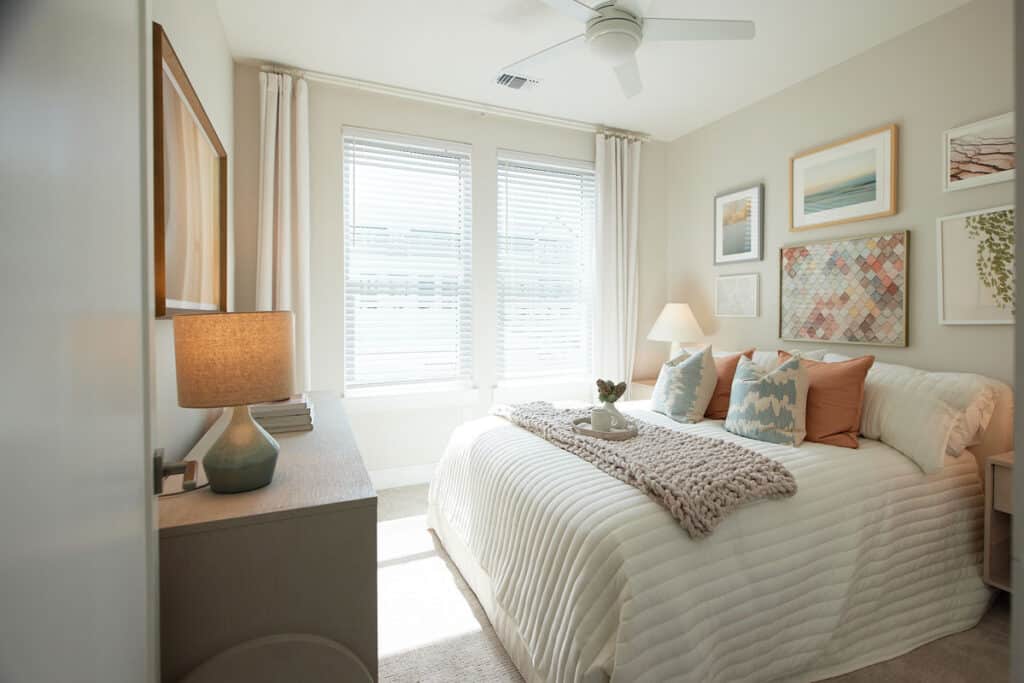 A modern bedroom with a neatly made bed, decorative pillows, wall art, a bedside lamp, a dresser with another lamp, and two large windows letting in natural light.