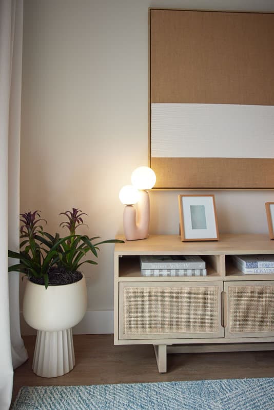A sideboard with rattan doors holds books, framed art, and a modern lamp with two round bulbs. A potted plant sits to the left, and a large abstract artwork hangs above.