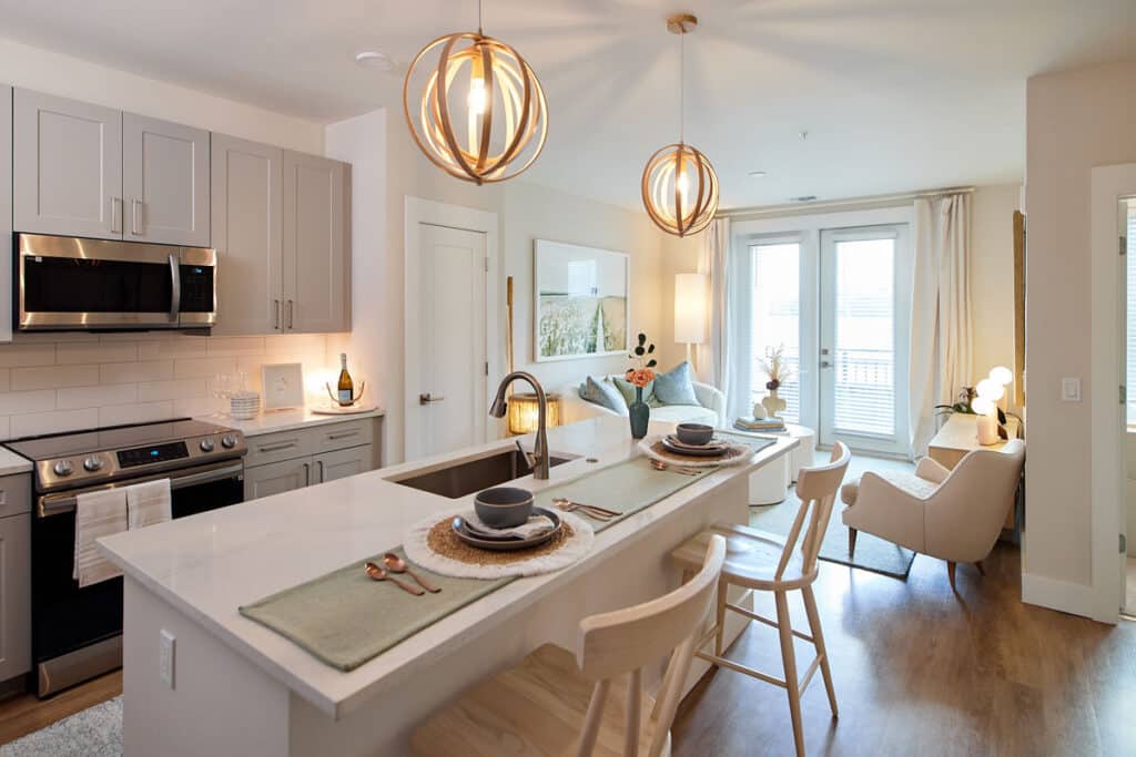 Modern kitchen and living area with light wood floors, gray cabinets, stainless steel appliances, pendant lighting, and a breakfast bar set for two.
