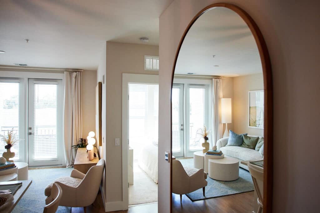 A modern living room and bedroom with neutral decor are visible through an arched mirror, featuring large windows, armchairs, a sofa, and soft natural light.