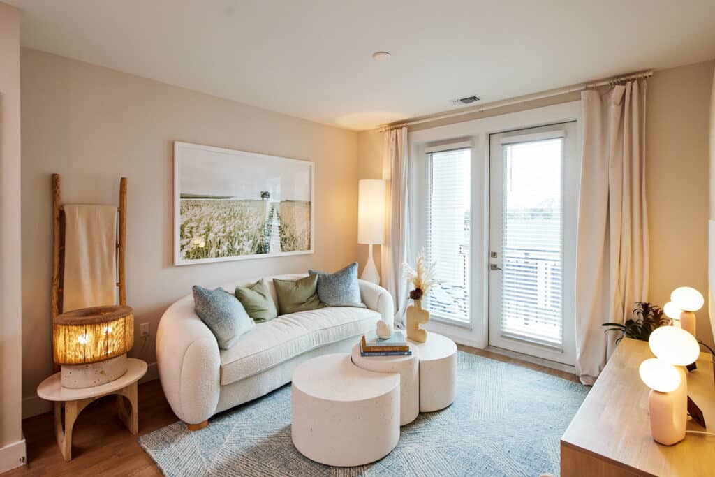 A modern living room with a white sofa, round coffee tables, a floor lamp, and large windows with blinds. Neutral tones and minimalist decor create a cozy atmosphere.