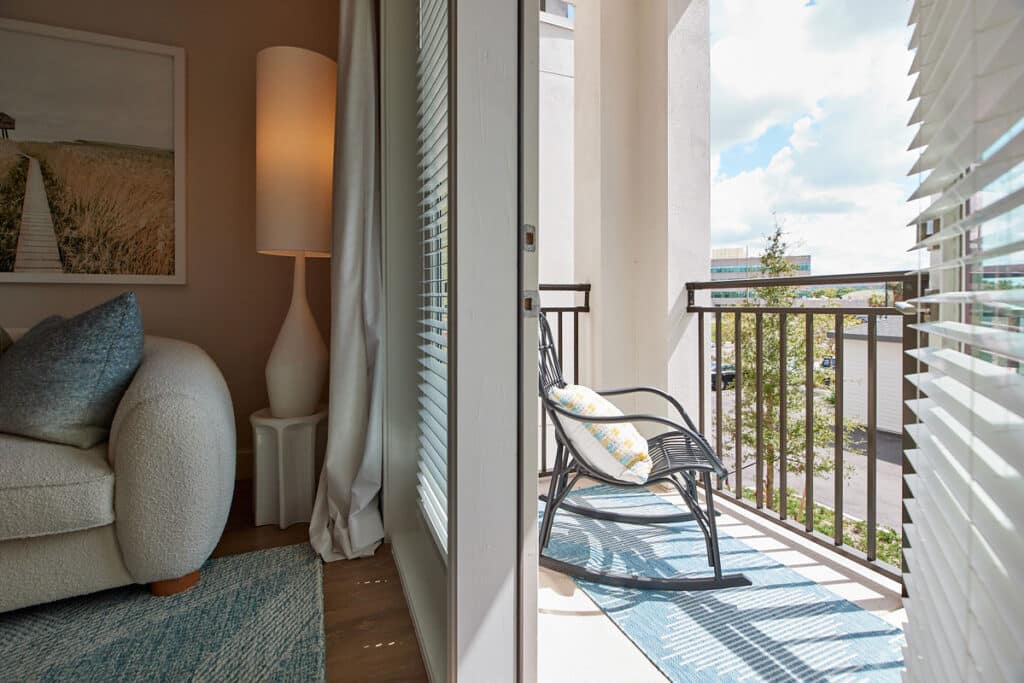 A bright room with a white couch and tall lamp opens onto a small balcony with a black rocking chair and pillow, overlooking a parking lot and trees.
