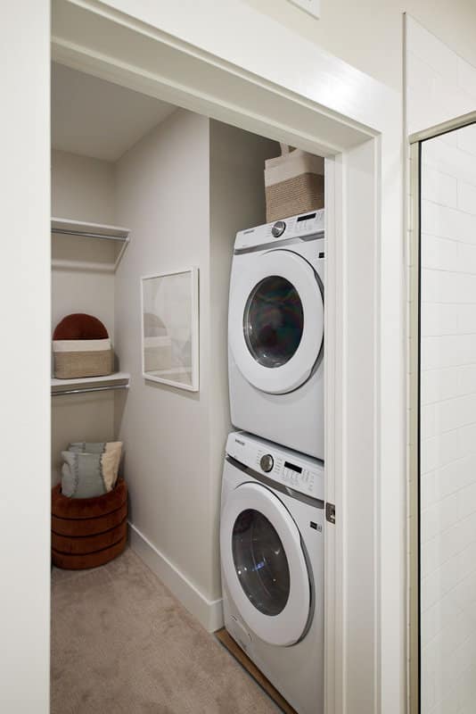 A compact laundry area with a stacked washer and dryer next to a small closet space with shelves, baskets, and decorative pillows.
