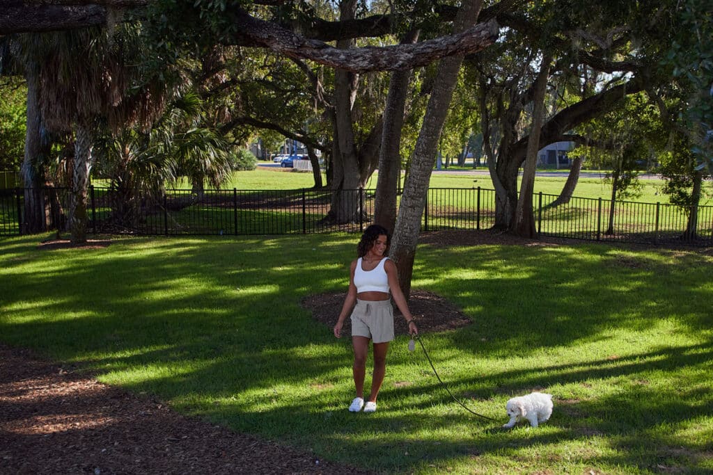 A woman walks a small white dog on a leash in a grassy park with large trees and a black metal fence in the background.