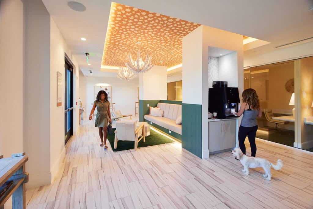 Two women are in a modern, well-lit lounge area; one is walking while the other uses a beverage machine, and a small white dog stands nearby on the tiled floor.