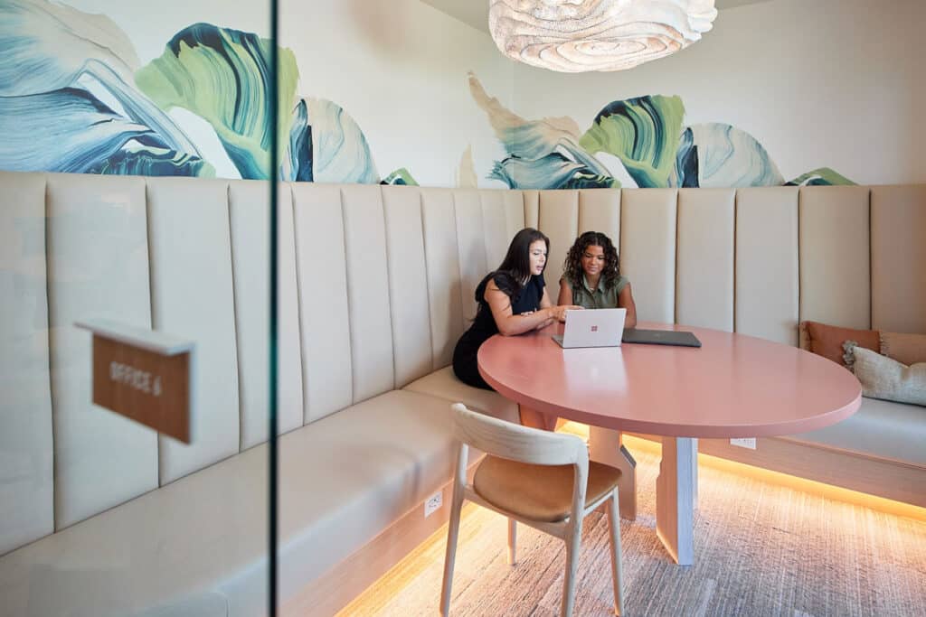 Two women sit at a round pink table in a modern office space, looking at a laptop together.