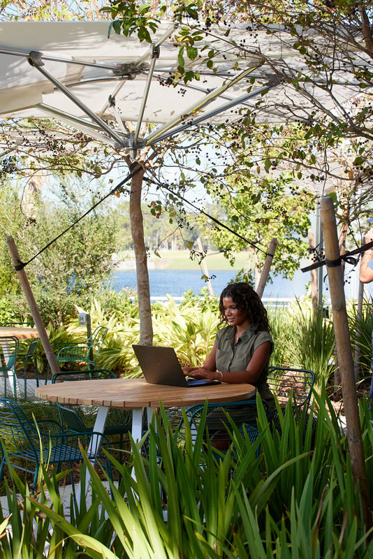 A woman sits at a round outdoor table working on a laptop, surrounded by greenery and trees, with a body of water visible in the background.