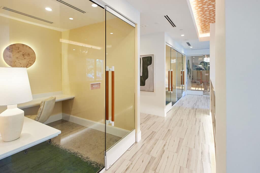 Modern office hallway with light wood flooring, glass-walled rooms, a desk with a lamp, contemporary art, and recessed lighting in the ceiling.