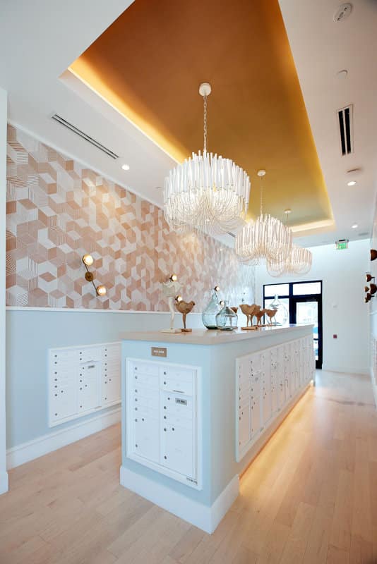 A modern mailroom with white mailboxes, geometric wallpaper, wooden floor, decorative glass and ceramic animals, and two large chandeliers hanging from a gold ceiling inset.