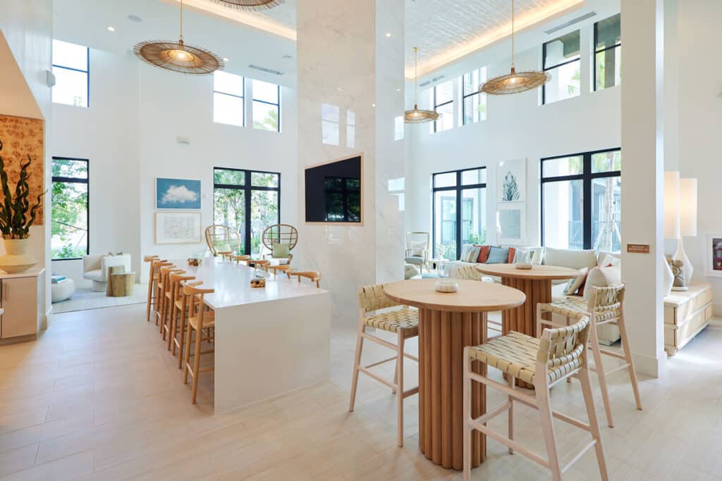 Modern open-concept living space with large windows, a central kitchen island, round tables with chairs, lounge seating, and light wood and white decor.