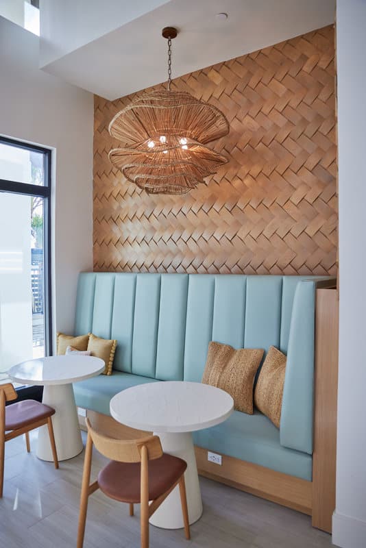 A cozy café seating area with light blue upholstered bench, two round white tables, wooden chairs, wicker pillows, woven wall decor, and a rattan pendant light.